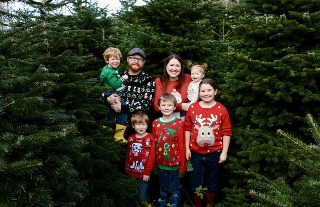 Slater family (two parents, three boys, two girls) wearing festive sweaters and standing amid fir trees.