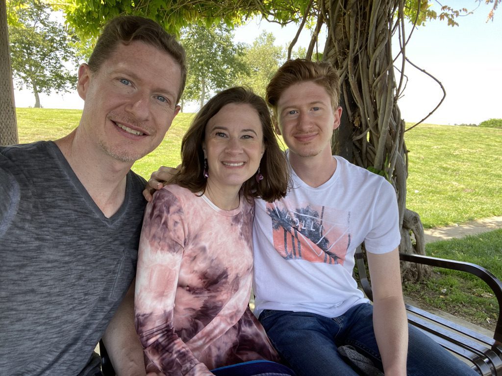 Selfie perspective of a man, woman, and son sitting on a park bench with a tree and grass in the background.
